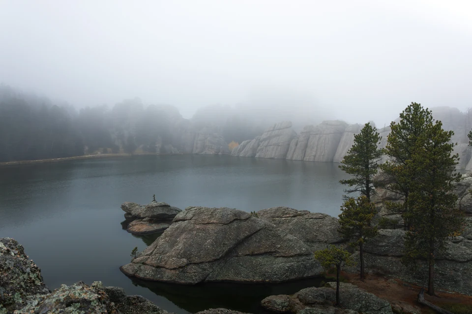 A still lake, granite boulders, and pine trees disappear into low fog.