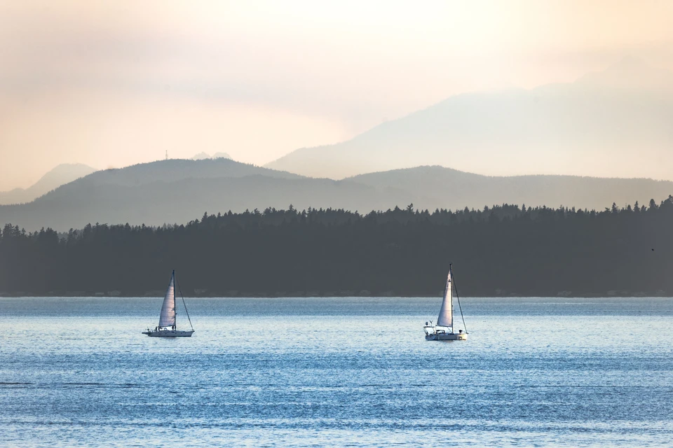 Two sailboats cross a blue inlet beneath layers of hazy mountains.