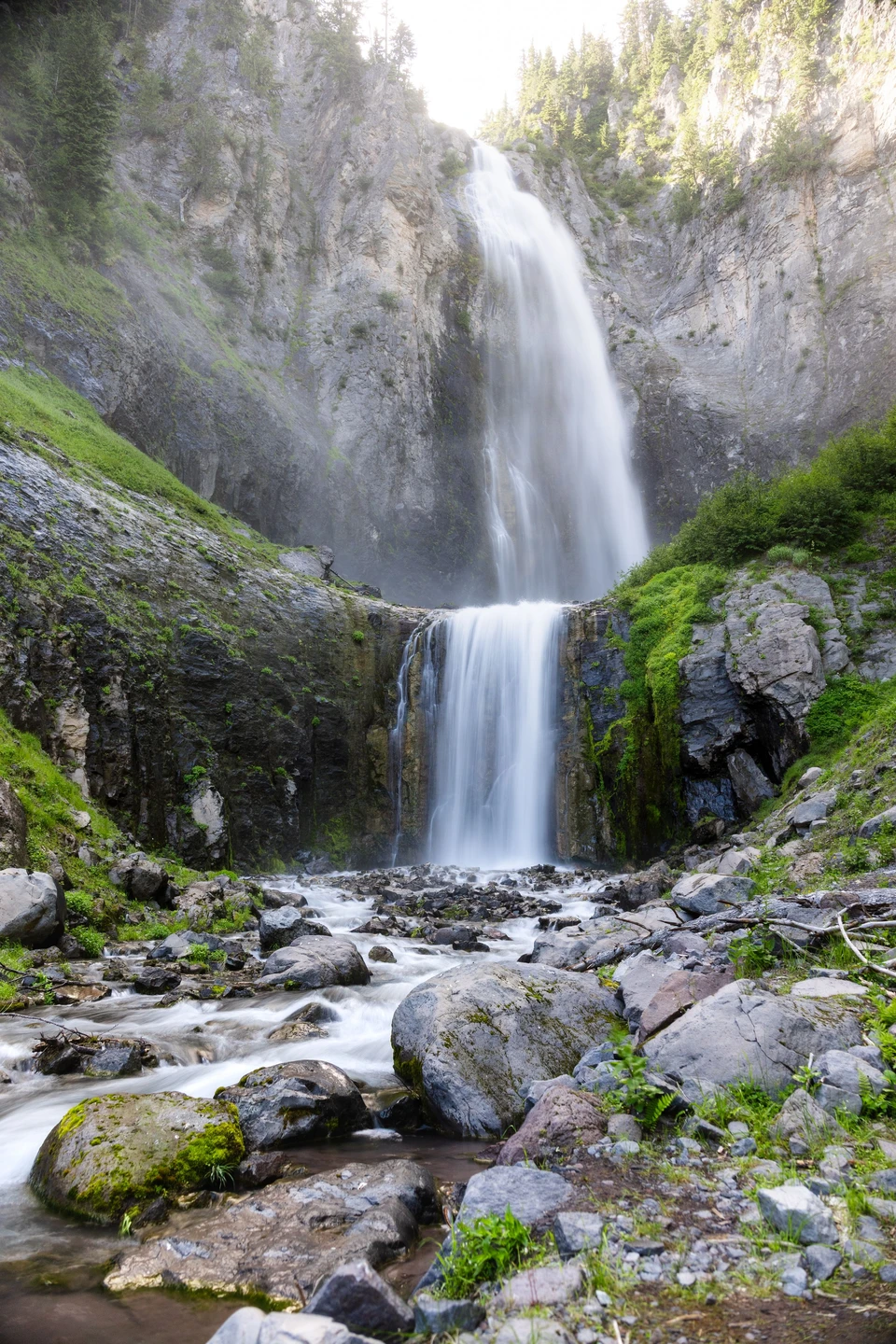 A two-tier waterfall flows over dark rock among moss and ferns.