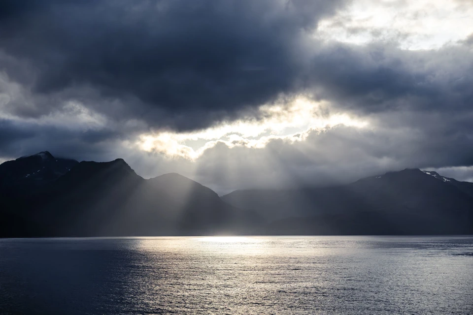Shafts of sunlight fall between storm clouds onto a mountain inlet.